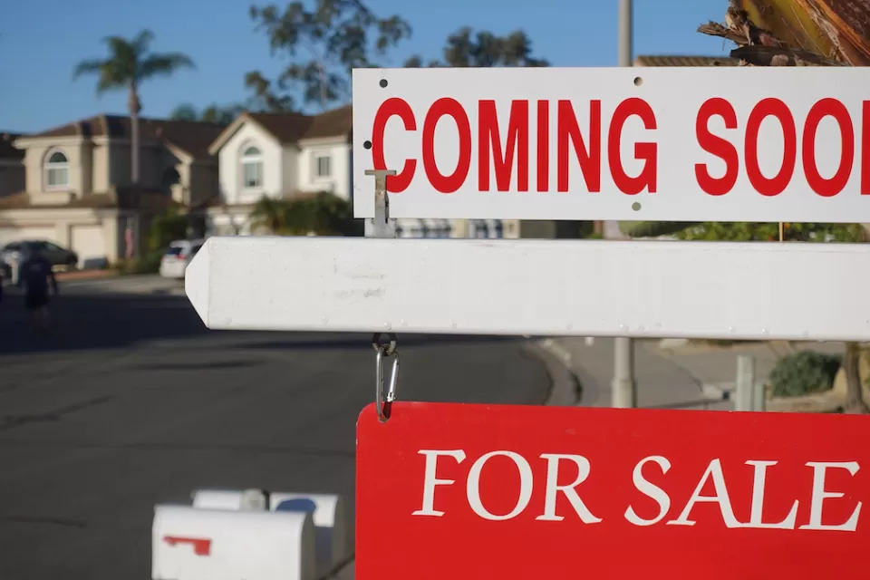 Close-up on red and white "Coming Soon" and "For Sale" signs in front of single-family homes on sunny day.